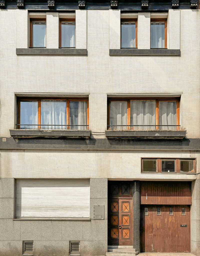 A mid-20th century building façade covered with small square mosaic tiles. The structure has three levels with simple rectangular windows framed in wood and concrete. The ground floor includes a wooden garage door, a main entrance door with geometric patterns, and a closed shutter. The overall look is plain yet balanced, with a touch of retro elegance.