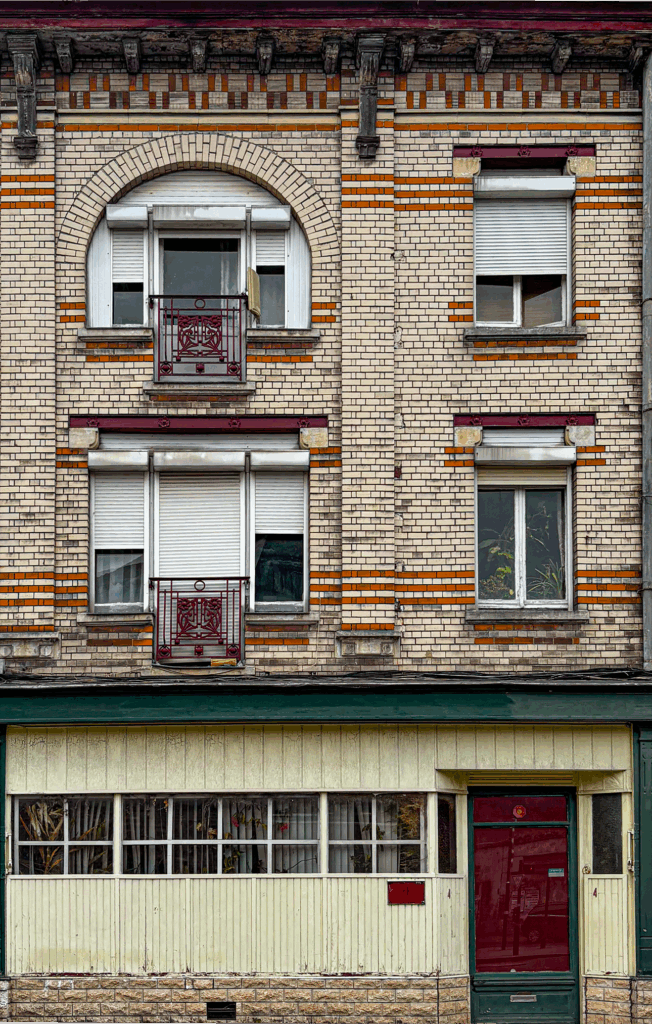 Immeuble résidentiel modeste du début du XXᵉ siècle, doté d’une façade en briques décoratives rouges et blanches. Les étages supérieurs présentent des fenêtres cintrées et rectangulaires équipées de volets roulants blancs et encadrées de motifs en briques, avec des balcons en fer forgé peints en rouge foncé. La corniche est soutenue par des consoles en bois sculptées, ajoutant une touche ornementale. Le rez-de-chaussée contraste par un lambris vertical clair, une rangée de fenêtres garnies de rideaux et une porte d’entrée rouge foncé. L’ensemble associe une conception fonctionnelle à de petites touches décoratives typiques de l’architecture urbaine ouvrière.
