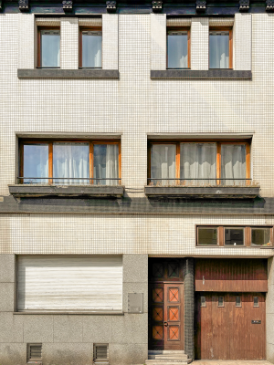 A mid-20th century building façade covered with small square mosaic tiles. The structure has three levels with simple rectangular windows framed in wood and concrete. The ground floor includes a wooden garage door, a main entrance door with geometric patterns, and a closed shutter. The overall look is plain yet balanced, with a touch of retro elegance.