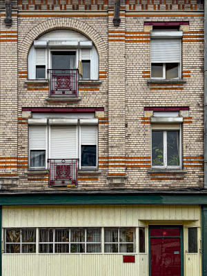 Mmodest early 20th-century residential building with a decorative brick façade in red and white. The upper floors feature arched and rectangular windows fitted with white roller shutters and framed by patterned brickwork, with wrought-iron balconies painted dark red. The cornice is supported by carved wooden brackets, adding ornamental detail. The ground floor contrasts with light vertical paneling, a row of windows with curtains, and a dark red entrance door. The overall appearance combines functional design with small decorative touches typical of working-class urban architecture.