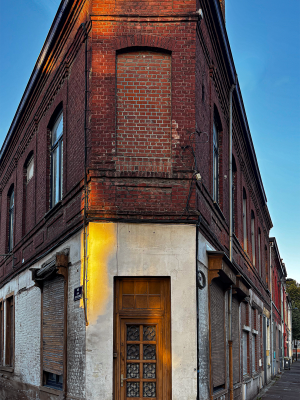 A building set at the corner of two streets, shaped like a triangle reminiscent of a “flatiron.” Its façade is made of dark brick, with several openings either bricked up or sealed with metal shutters. On the ground floor, a glass-panelled door with decorative details stands at the pointed tip of the building, framed by a lighter plaster section contrasting with the brick. Electrical cables run across the façade, and a streetlamp juts out above. On the sidewalk, trash bins and a traffic sign complete the scene. The overall impression is one of abandonment, a place where time seems to have stopped.